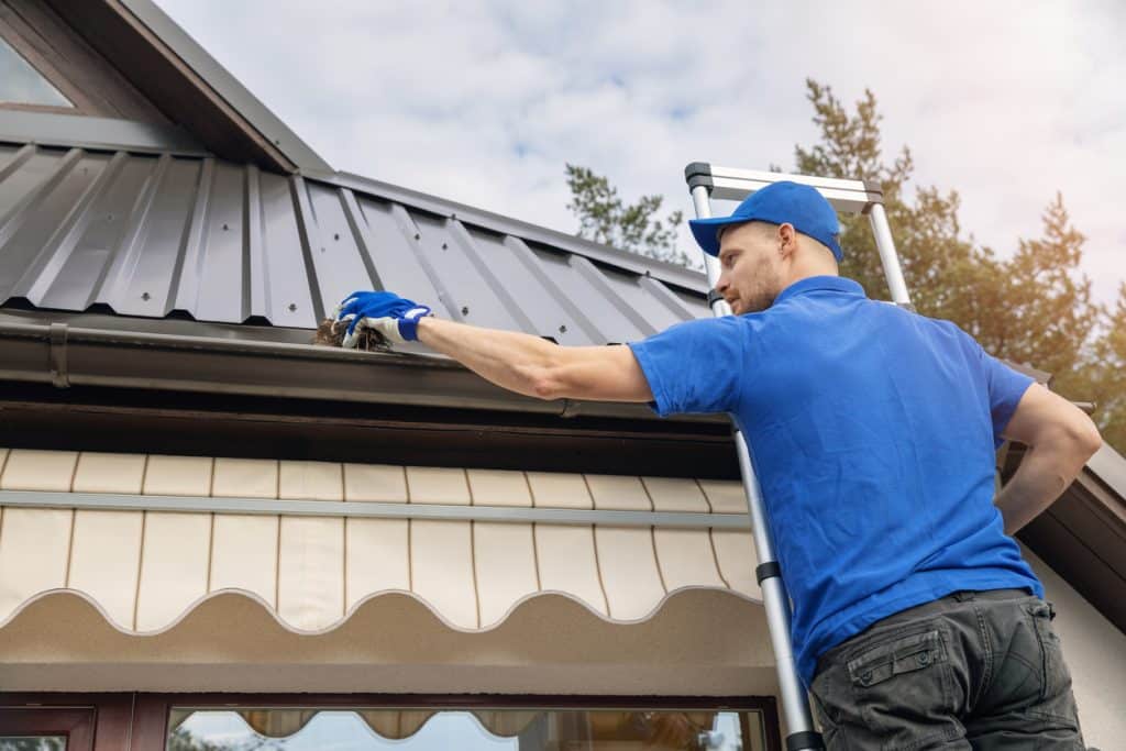 A worker in a blue uniform and cap uses a ladder to clean debris from a residential roof gutter.