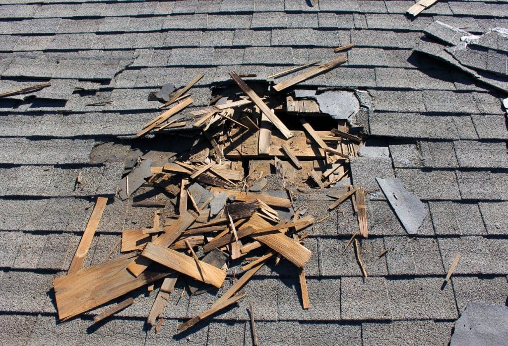 Close-up of a residential roof with severely damaged asphalt shingles, a large hole exposing the underlying wooden roof structure, and broken wood fragments scattered across the roof surface.
